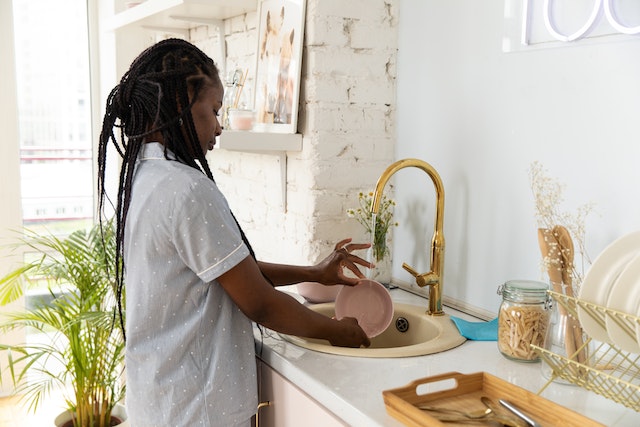 a tenant with box braids washes their dishes in a kitchen sink set into a white marble countertop with a gold faucet